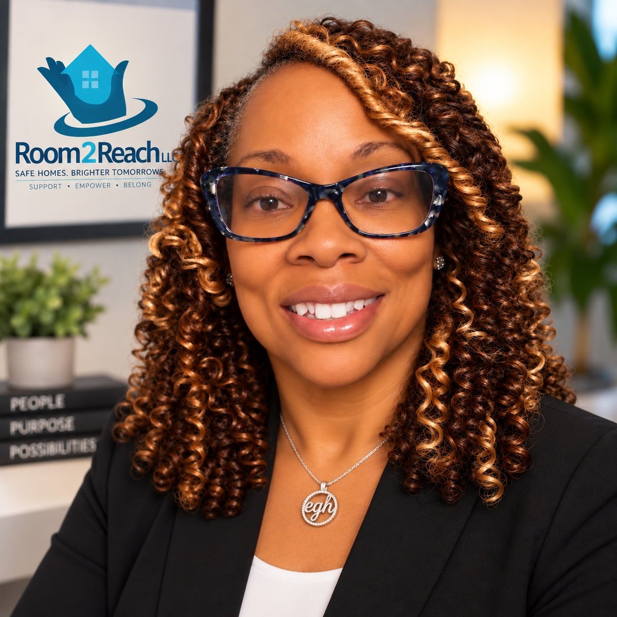 Professional woman with curly reddish-brown hair and glasses smiling at camera in front of Room2Reach logo